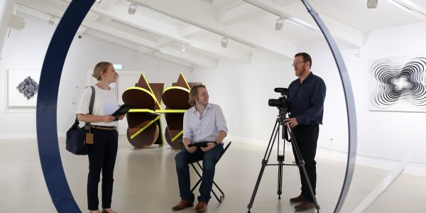 Three people stand in a museum space next to a video camera and an exhibit