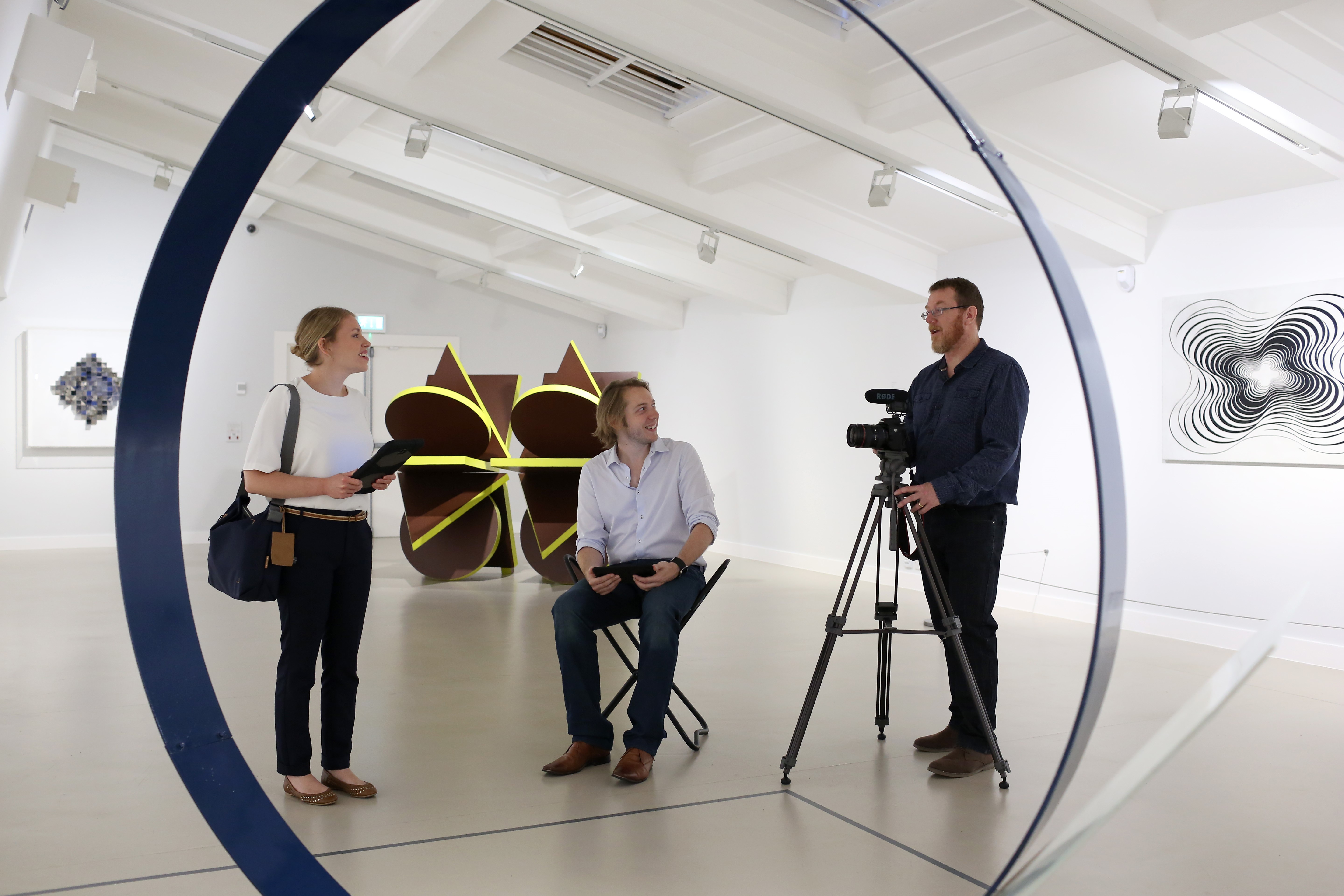 Three people stand in a museum space next to a video camera and an exhibit