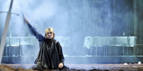 A man holds his hand up on a muddy stage, with a sword in the foreground.