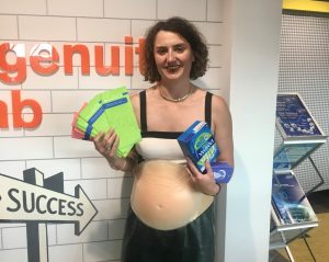 Olivia wearing a pregnancy belly standing in the Ingenuity Lab holding some leaflets and tampons 