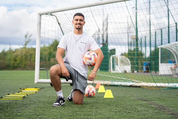 Daniel Damasceno de Faria kneeling on grass in front of a goal post holding a football