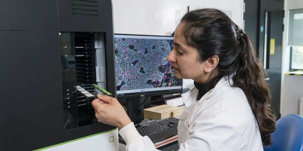 Sunny Sunshine with her dark brown hair tied back, wearing a white lab coat and holding samples in a black machine