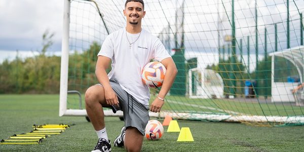 Daniel Damasceno de Faria kneeling on grass in front of a goal post holding a football