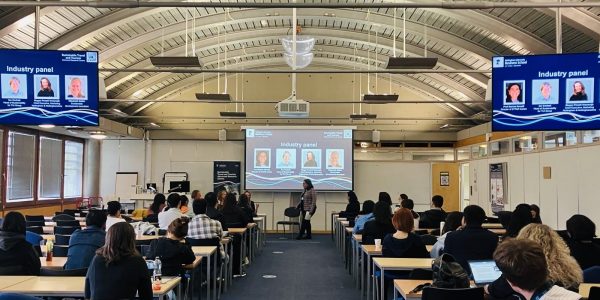 Marina Novelli at the front of a large lecture theatre for World Tourism Day 2025 with rows of students sat at tables facing the front with large screens at the front and sides