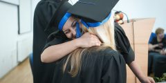 Two students wearing graduation gowns and caps embrace in a hug. The girl facing the camera with dark hair is smiling.