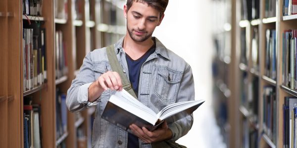 Male student in library surrounded by books is facing towards the camera whilst looking at an open book