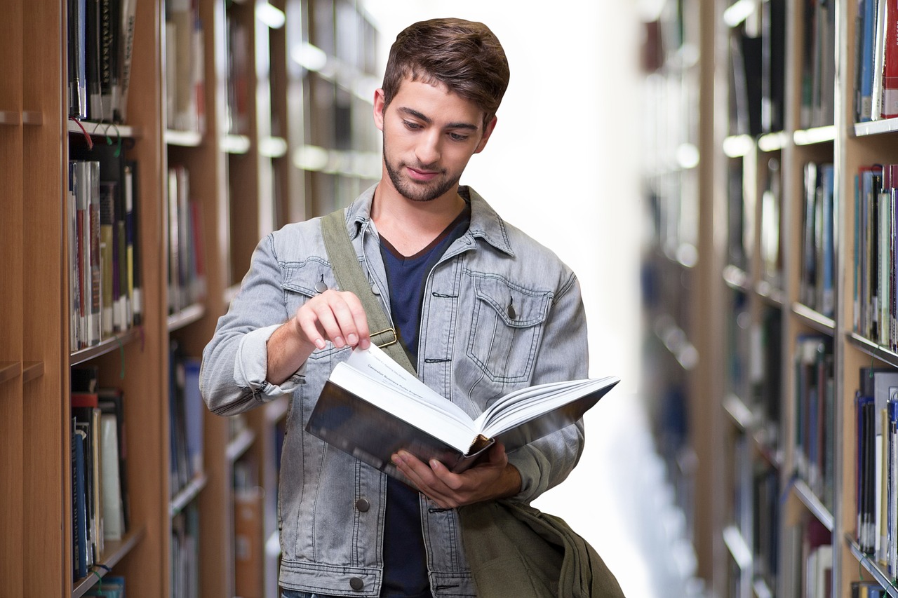 Male student in library surrounded by books is facing towards the camera whilst looking at an open book