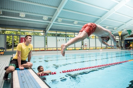 A male in red swimming trunks diving into a swimming pool with two other males watching