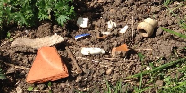 Photograph of pieces of pottery in a flower bed. Including a piece of flower pot, a stoneware bottle neck, some bent and rusty nails and a small headless china doll
