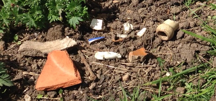 Photograph of pieces of pottery in a flower bed. Including a piece of flower pot, a stoneware bottle neck, some bent and rusty nails and a small headless china doll