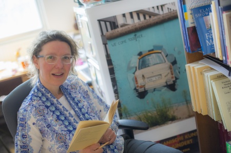 Heike sits in a chair with a book open in front of her, but looking at the camera. She is dressed in a blue shawl and has clear-framed glasses on