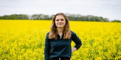 Lisa, a young woman in a green top, is standing in a field of yellow flowering oilseed rape