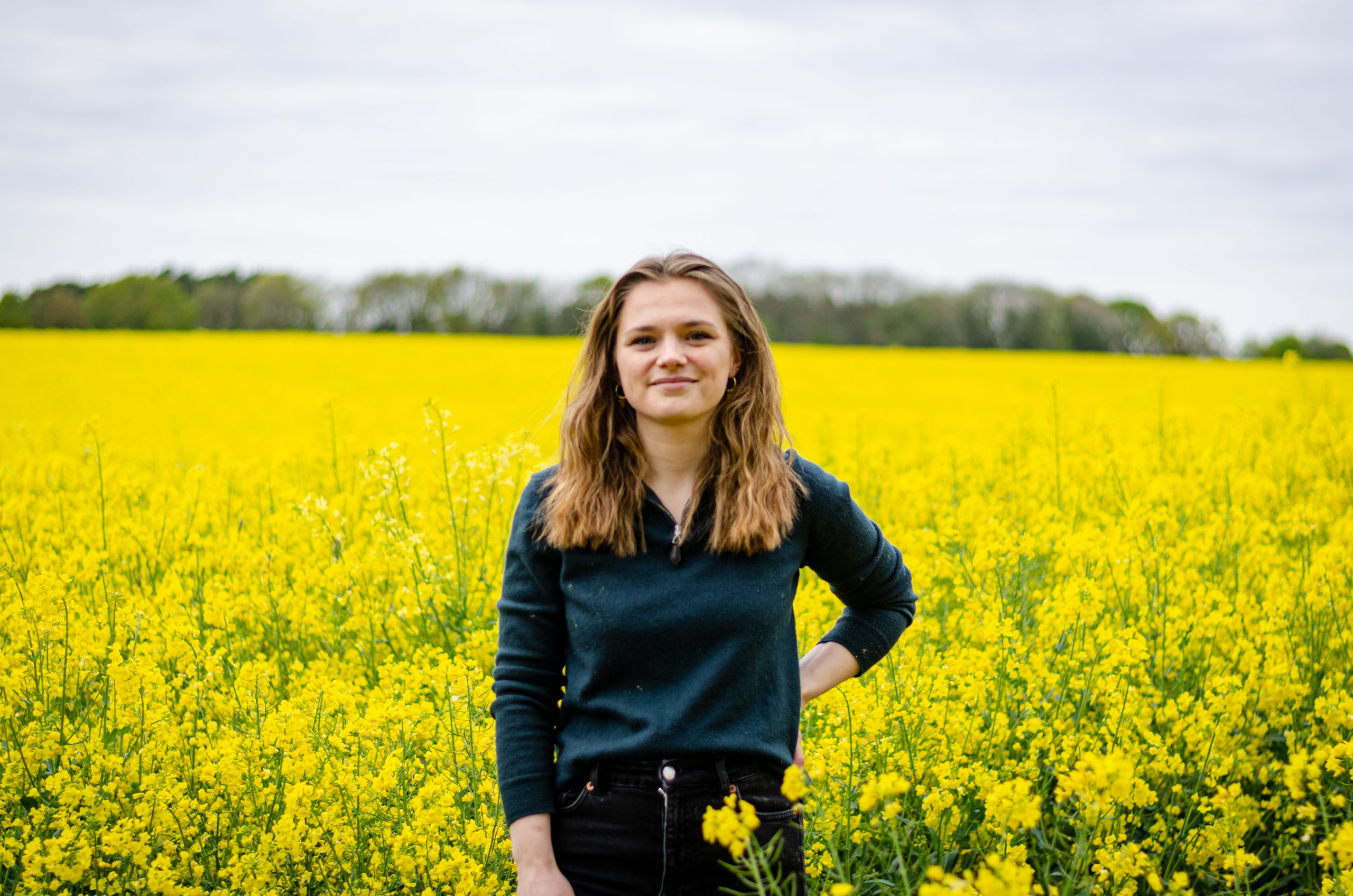 Lisa, a young woman in a green top, is standing in a field of yellow flowering oilseed rape