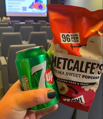 Close up of a hand holding can of soda and a bag of popcorn with the seats and screen of Hallward Library screening room in the background