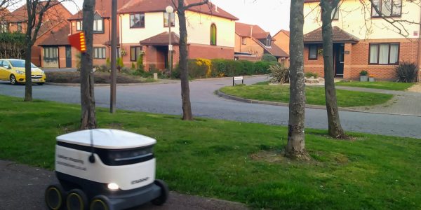 'Starship' delivery robot on a pavement in Milton Keynes with some trees and suburban houses in the background