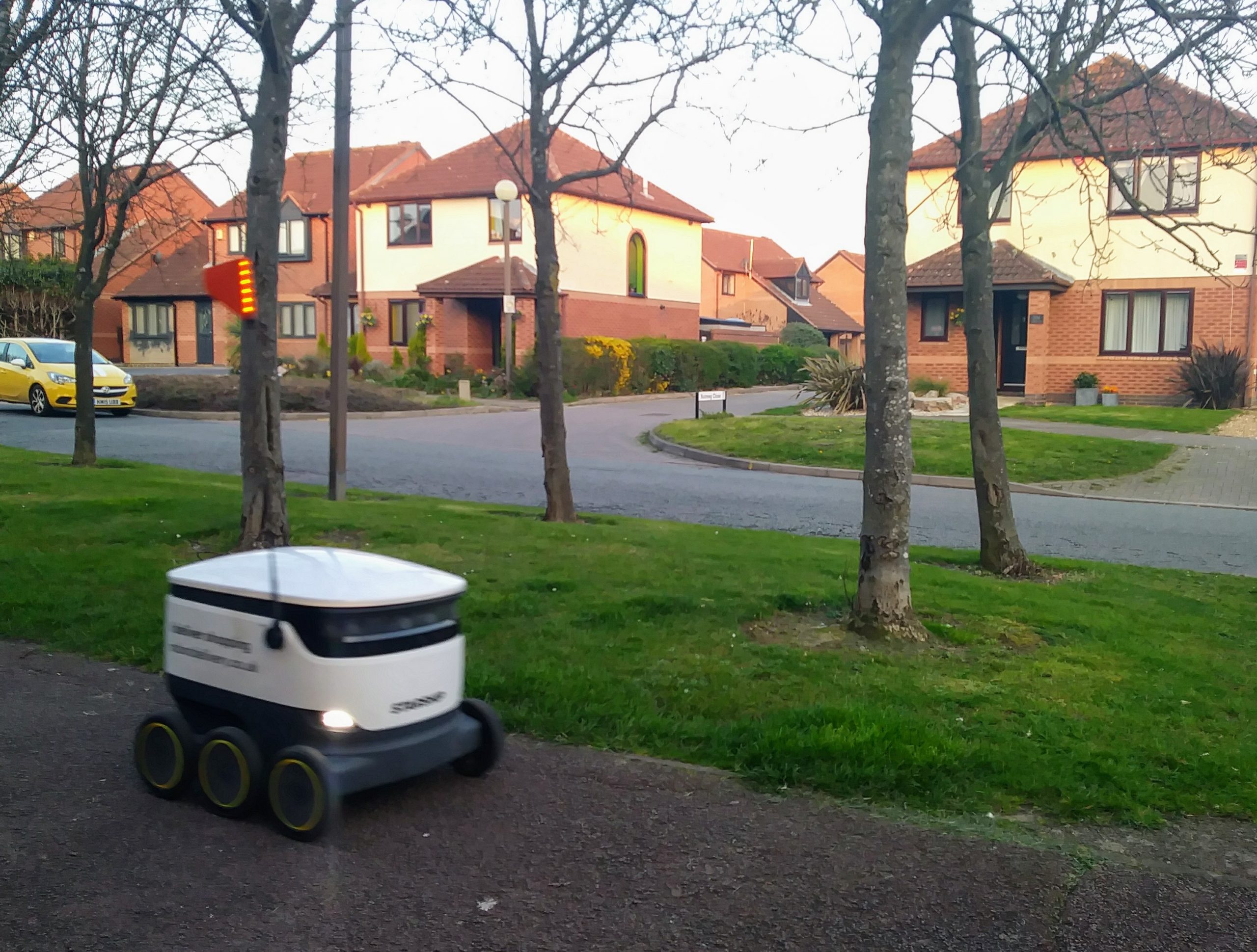 'Starship' delivery robot on a pavement in Milton Keynes with some trees and suburban houses in the background
