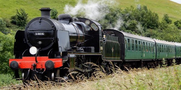 Black steam locomotive with green carriages going past green hills (Corfe Castle)
