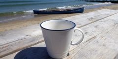 white cup on wooden bench overlooking blue see with a canoe in the foreground
