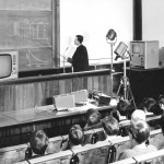 Shows the lecturer, dressed in a black gown, pointing at a diagram on the blackboard; also shows various electrical equipment, and the first 3 rows of students.