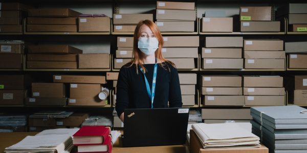 Young woman wearing a mask looking at the camera, standing in front of shelves of archive boxes, next to a table on which is a laptop, archive documents and books.
