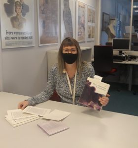 Woman wearing a face mask sitting at a desk with unbound chapters from the book in a box novel The Unfortunates in front of her and in her hands