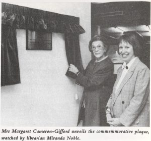 Black and white photograph of two women standing next to a plaque which has been unveiled from behind curtains. The caption reads, "Mrs Margaret Cameron-Gifford unveils the commemorative plaque, watched by librarian Miranda Noble".