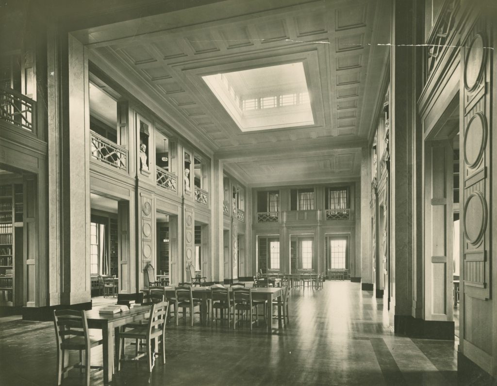Photograph of the University Library in the Trent Building, around 1928. A tall ornate room with large windows, tables and seats, with bookcases at the edge of the room.