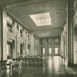 Photograph of the University Library in the Trent Building, around 1928. A tall ornate room with large windows, tables and seats, with bookcases at the edge of the room.