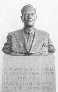 Black and white professional photograph of the head and shoulders bronze bust of Bertrand Leslie Hallward, first vice-chancellor of the University of Nottingham, 1948-1965; statue is mounted on a wooden plinth engraved with the above information, along with the words 'collegium invenit, reliquit universitatem'.