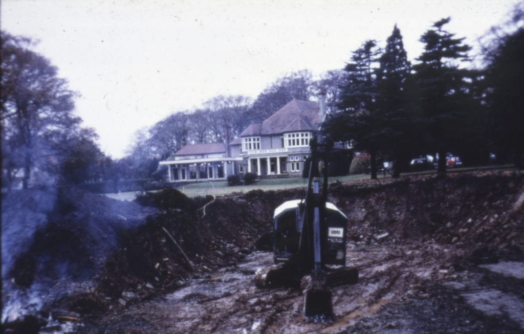 Photograph showing a mechanical digger in a mud trench in the grounds of Lenton Mount house.
