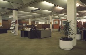 Photograph of the interior of a library, with a man sat behind a desk in the middle of a large open plan space with checkered carpet, white pillars, and pot plants.