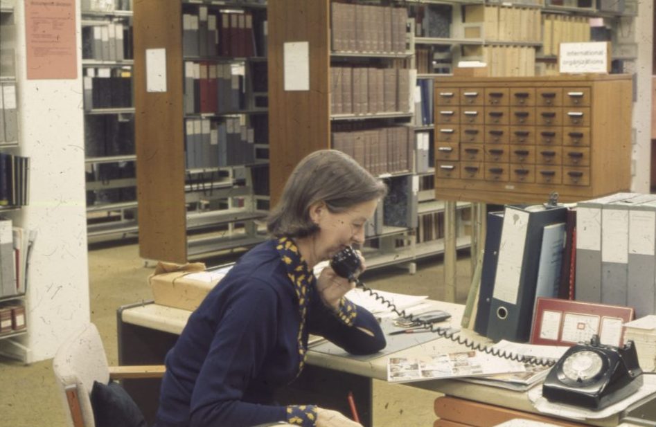 Photograph of female library staff member at an enquiry desk answering a corded telephone. Behind her are shelves of books and a card catalogue made up of many small wooden drawers.