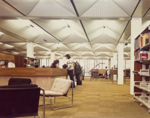 Photograph showing the inside of the library, with readers stood at a help desk with a staff member behind it, high ceilings with bright lights, a seating area, and shelves with books.
