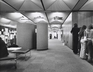 Black and white photograph showing the interior of the library with high ceilings, bright lights, and students gathered around. There are also some seats and noise cancelling dividers next to a pinboard full of flyers.