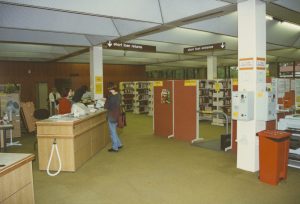 Photograph of sectioned off part of library, showing gates with stacks of books beyond them. There is also a help desk with a library staff member and a student.