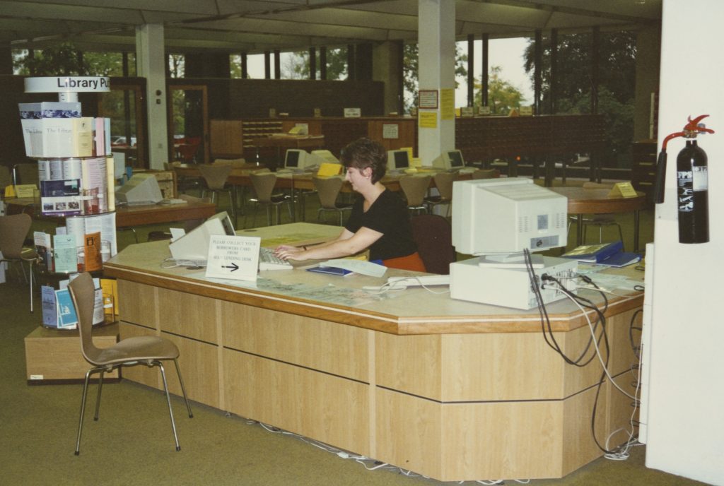 Photograph of female library staff member sat behind counter. There is a large, bulky PC on the desk with trailing cables. Next to the desk is a revolving leaflet stand.