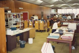 Photograph showing staff working in the library at desks with a mixture of computers and typewriters. There are trolleys of books and shelving racks.
