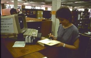 Photograph of woman scanning a barcode within an open library book using a pen-like device with a cable. On the desk in front of her is a 1980s computer, as well as ink stamps.