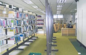 Photograph showing shelving and unfinished shelving being assembled. In the background you can see book trolleys and a library lending desk.