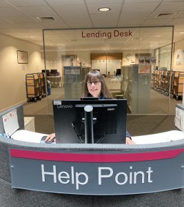 Photograph of Tracey sat behind a desk with a sign saying "HELP POINT"