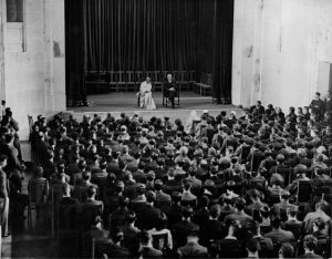 Black and white photograph of a large room. Two people are sat on chairs on the stage in front of a crowd of seated students.