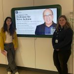 Two female students standing by a screen promoting Professor Sir Steve Jackson's lecture.