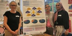 Three women smiling next to a poster about eczema and a table with fruit, pens and paper on it.