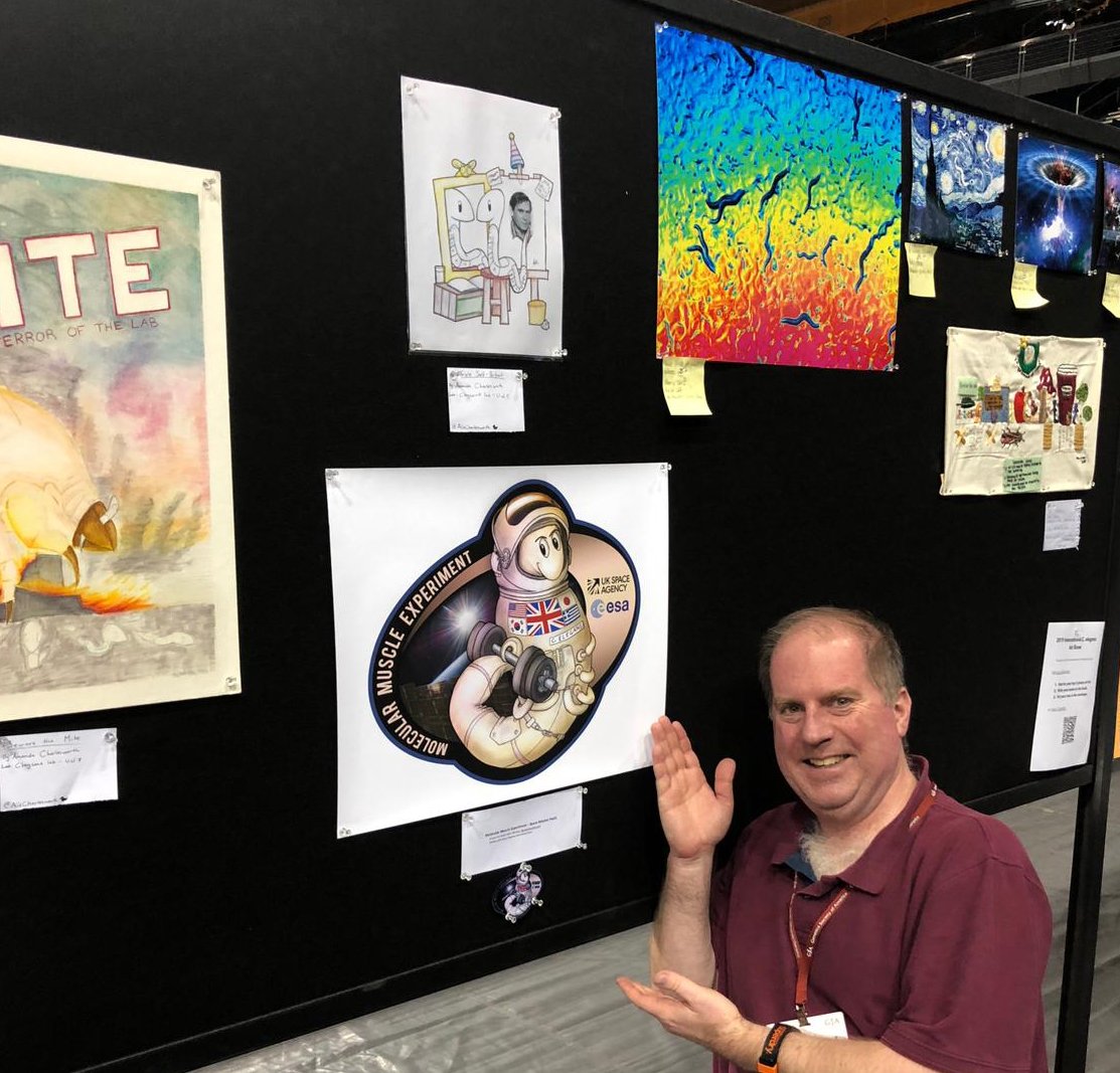 Professor Nate Szewczyk gesturing towards a drawn Worms in Space patch on a wall of artwork
