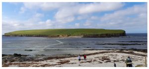 Photo of the Brough of Birsay, a small, round, and grass covered island. In the foreground is a narrow stretch of sea that becomes a causeway at low tide.