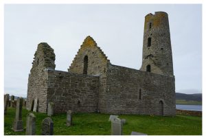 Photo of St Magnus's church, built out of stone, the diminutive, roofless church has a round tower at one end. Located on the water's edge with grave stones in the foreground.