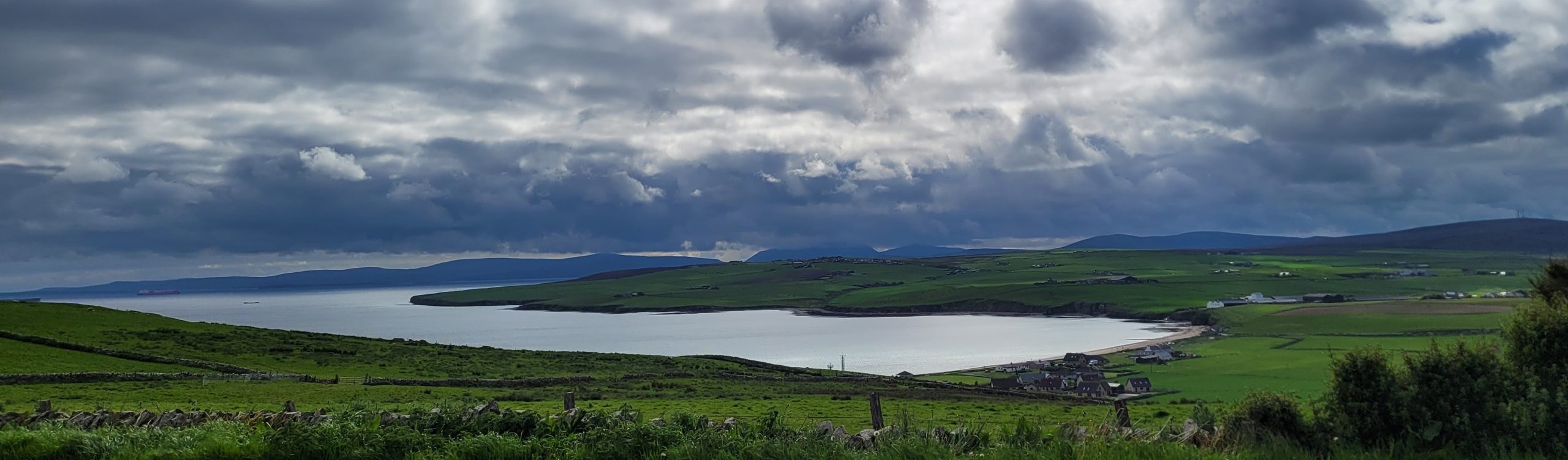 Photograph taken looking down to the bay of Scapa, a deeply curved bay, surrounded by farm land.