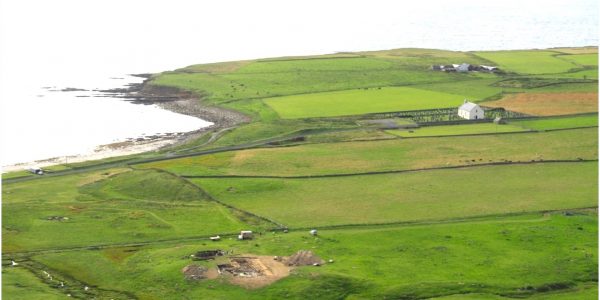 Colour photograph showing green fields along the Bay of Skaill coastline of the Orkney mainland. In the middle ground is distinct mound and archaeological dig site.