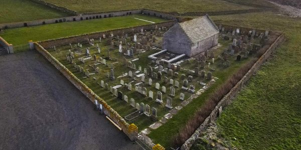 Aerial colour photograph of St Boniface church. A small stone building lying in a grassy walled graveyard in fields leading down to the sea.