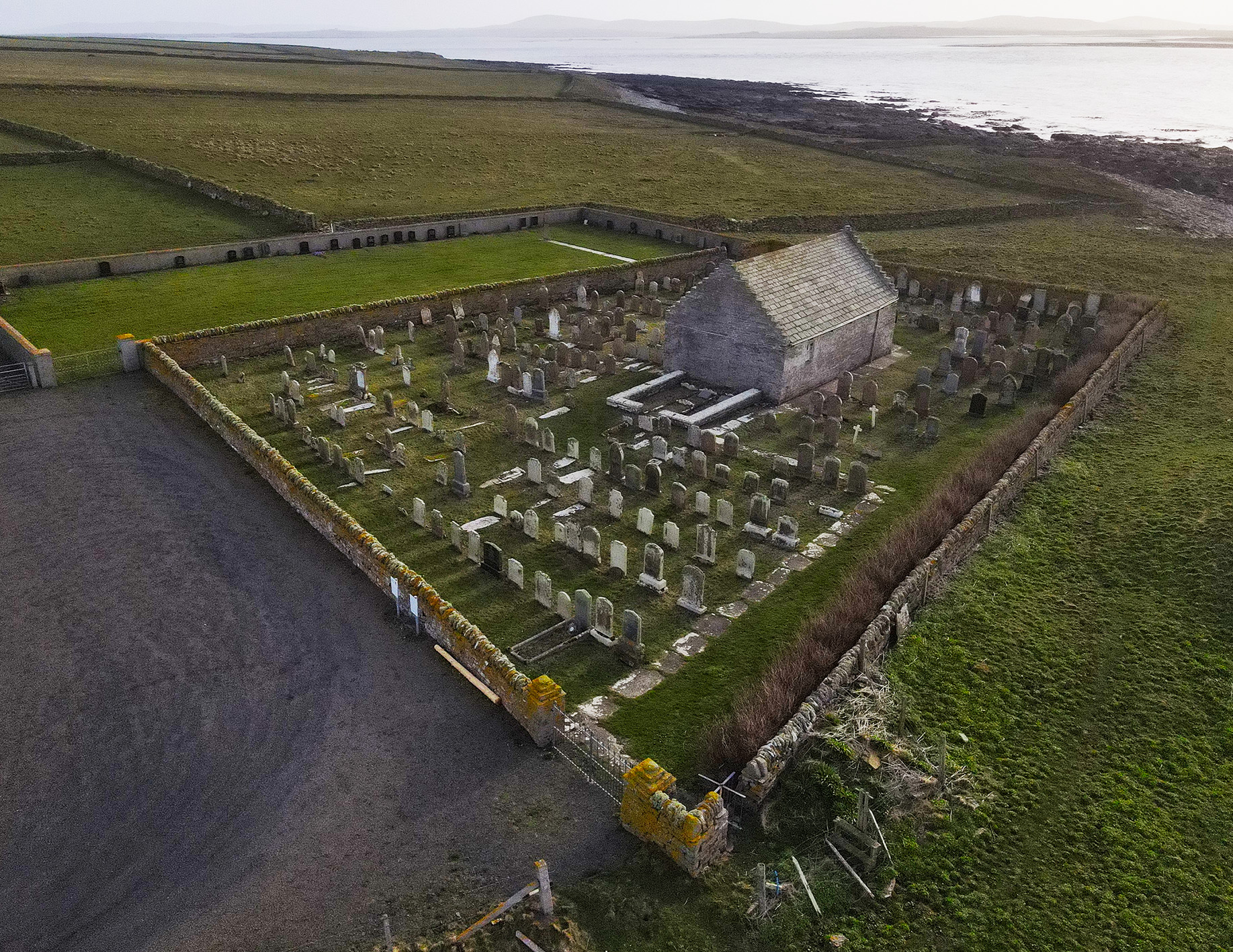 Aerial colour photograph of St Boniface church. A small stone building lying in a grassy walled graveyard in fields leading down to the sea.